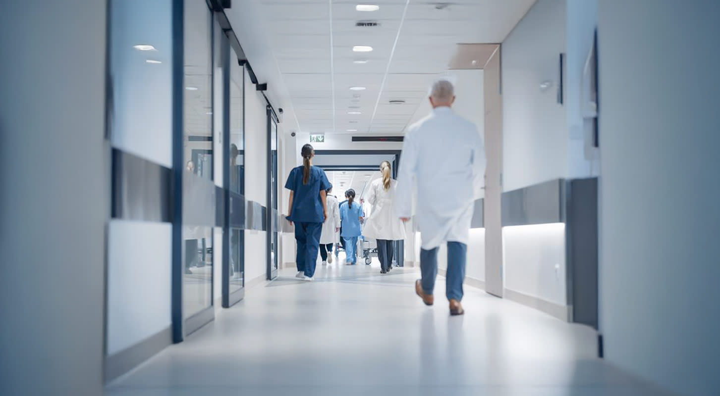View down a bright, sterile hospital hallway with multiple healthcare workers in white coats and scrubs walking away from the camera.